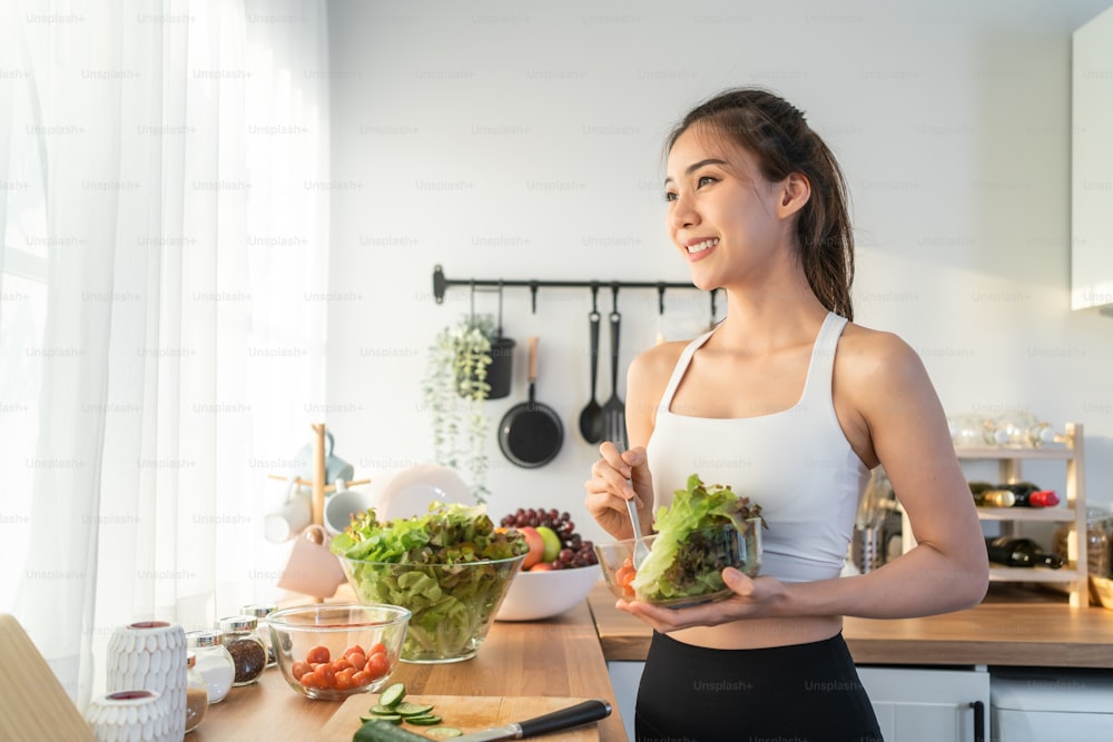 Asian attractive sport woman holding salad bowl and eat vegetables. Beautiful sport girl in sportswear enjoy eat clean vegetables after exercise for health in house. Diet and Healthy food concept.