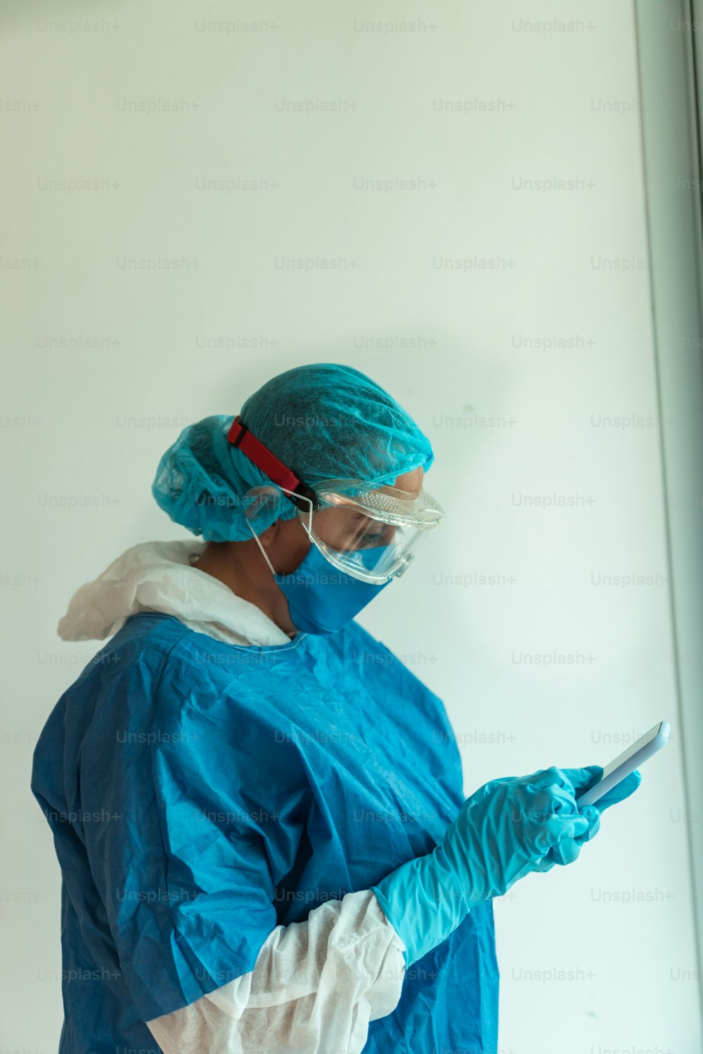 Médico joven y serio vestido con un uniforme azul y una máscara médica trabajando con un teléfono celular y guantes de látex en una clínica