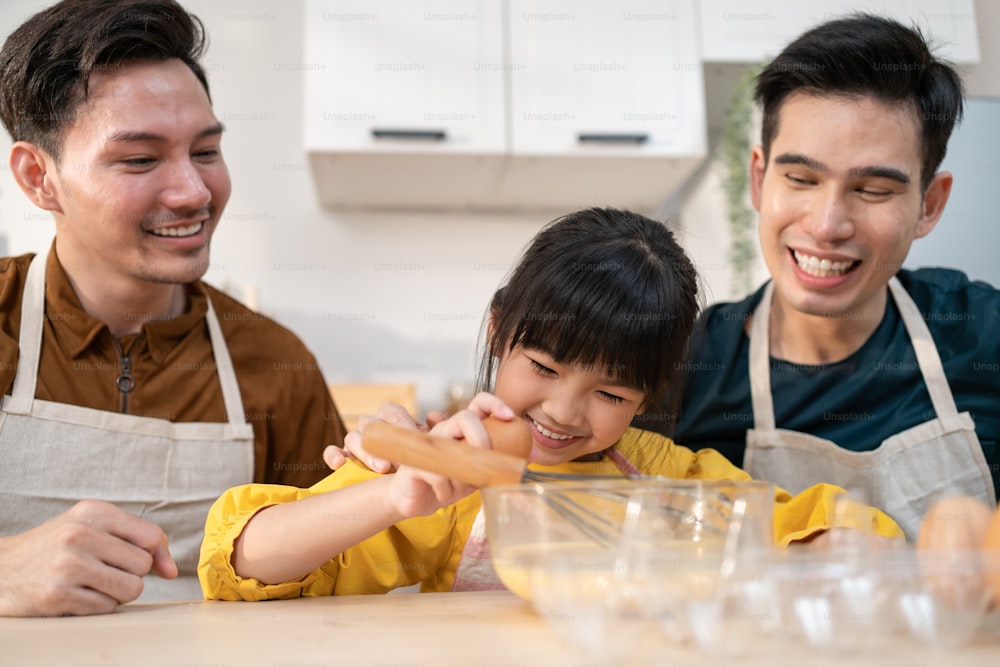 Asian attractive LGBTQ gay family teach girl kid making yeast dough. Handsome male couple take care and spend time with little adorable child bake bakery in kitchen, enjoy parenting activity at home.