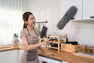Servizio di pulizia asiatico donna che pulisce la stanza della cucina a casa. La bella giovane donna delle pulizie indossa il grembiule e usa lo spolverino di piume per pulire il bancone sporco per le faccende domestiche o le faccende domestiche.