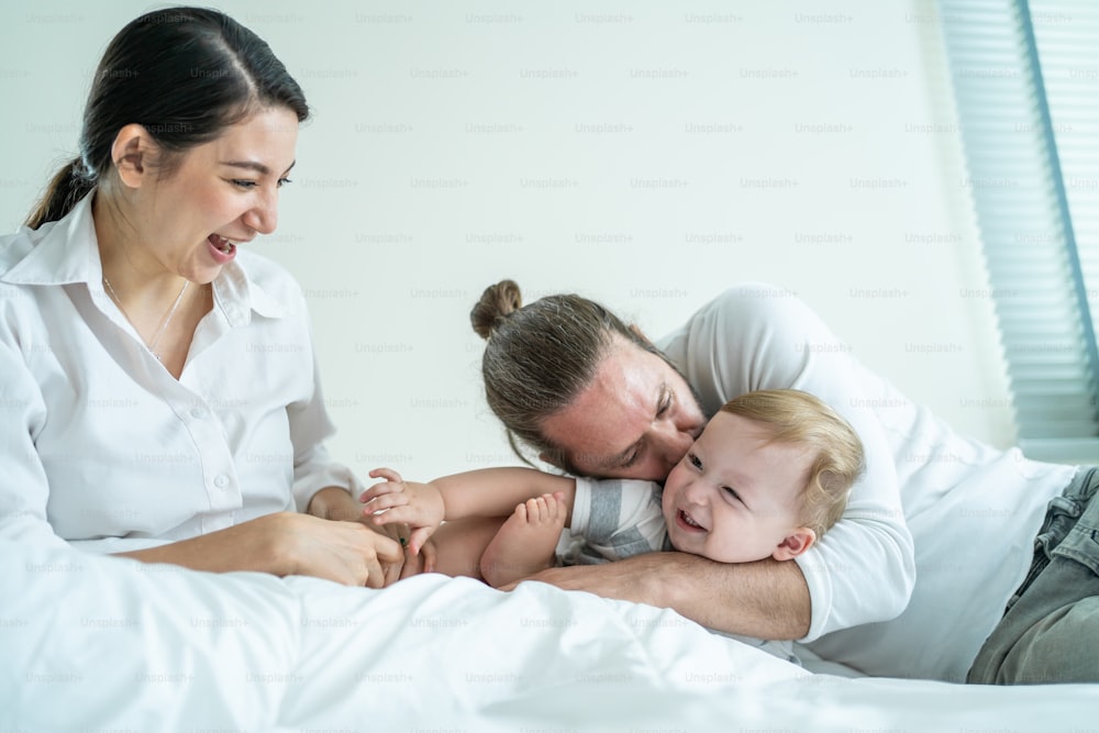 Caucasian parents playing with cute baby boy child on bed in bedroom. Happy family, attractive beautiful caring young mother and father feel happy looking and hug toddler kid son in morning in house.