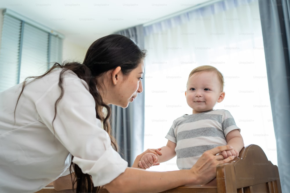 Caucasian mother play with cute baby boy child on infant bed at home. Happy family, attractive young woman mom talking to toddler son with happiness enjoy after wake up activity in bedroom in house.