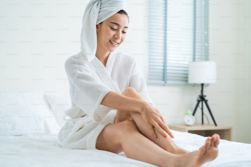 Une belle femme asiatique a mis des soins de la peau lisses et doux sur les jambes dans la chambre à coucher. Une jolie fille touche son genou et applique une lotion crème pour le rajeunissement avec douceur. Concept de beauté, de soins de la peau et de santé.