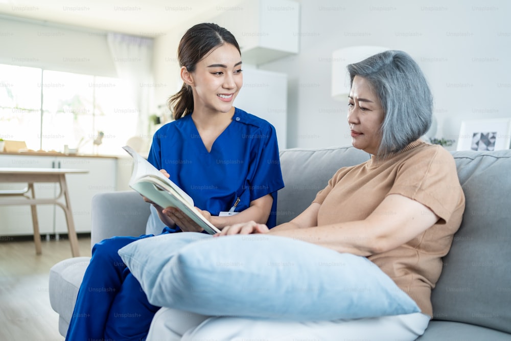 Asian woman nurse taking care of senior elderly female at nursing home. Young Caregiver Therapist doctor sit on sofa, read a book to happy older handicapped patient. Medical insurance service concept.
