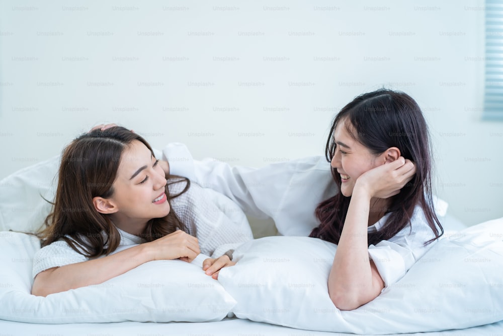 Asian beautiful lesbian couple lying on bed and looking at each other. Attractive romantic girl friend in pajamas spending morning leisure time together in bedroom in house. Homosexual-LGBTQ concept.