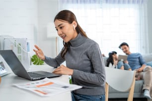 Asian businesswoman mother sit on table in living room, work from home. Attractive mom use laptop computer for online meeting in room with young kid daughter play with father on sofa together in house