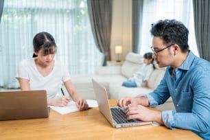 Asiatisch beschäftigt Eltern arbeiten aufgrund des Covid-19-Lockdowns von zu Hause aus, während Kinder dahinter fernsehen. Geschäftsvater und Mutter, die Laptop tippten und auf Kopierbuch schrieben, achteten nicht auf die kleine Tochter.