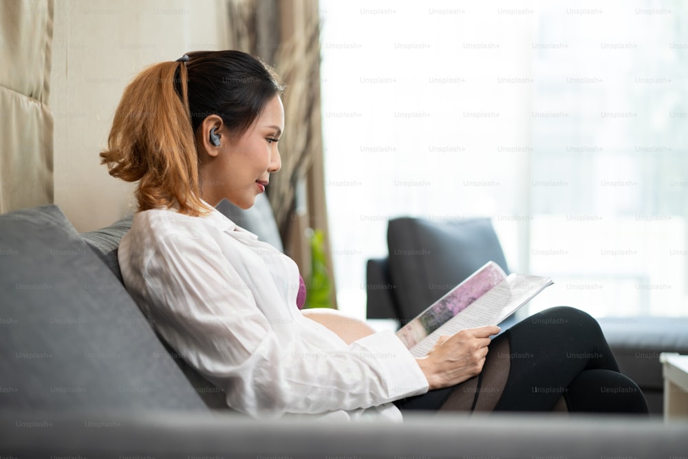 Asian young woman pregnant sitting and reading book with smile on sofa. Attractive female pregnancy sit on couch looking guidebook preparing for baby in womb, with happiness and smiling in living room