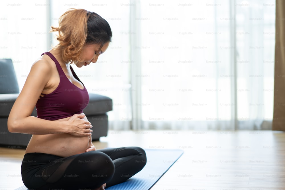 Asian young beautiful woman pregnant doing Yoga Pilates workout at home. Attractive female pregnancy in sportswear feel happy and relax, follow exercise instruction from online trainer in living room.