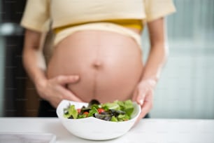 アジアの女性の妊娠中に立って野菜サラダを保持している接写。魅力的な美しい若い女性の妊娠スタンドと子宮内の赤ちゃんに触れ、キッチンで健康的な食べ物やベジタリアン料理を選びます。