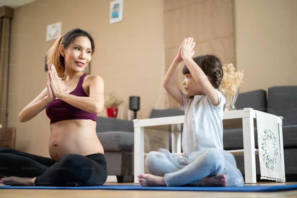 Asian young woman pregnant and little girl doing Yoga Pilates at home. Attractive mother pregnancy with kid daughter in sportswear feel happy and relax, follow exercise instruction from online trainer