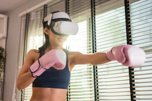 Sportiva asiatica che utilizza occhiali VR di tecnologia innovativa per l'esercizio. Bella ragazza attraente che indossa cuffie per realtà virtuale e tiene in mano il controller di gioco mentre fa allenamento di combattimento di boxe a casa.