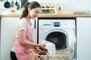 Asian beautiful woman put dirty clothes to washing machine in house. Attractive girl wear apron sit on floor, feel happy to loads laundry in washer appliance at home. Domestic-House keeping concept.