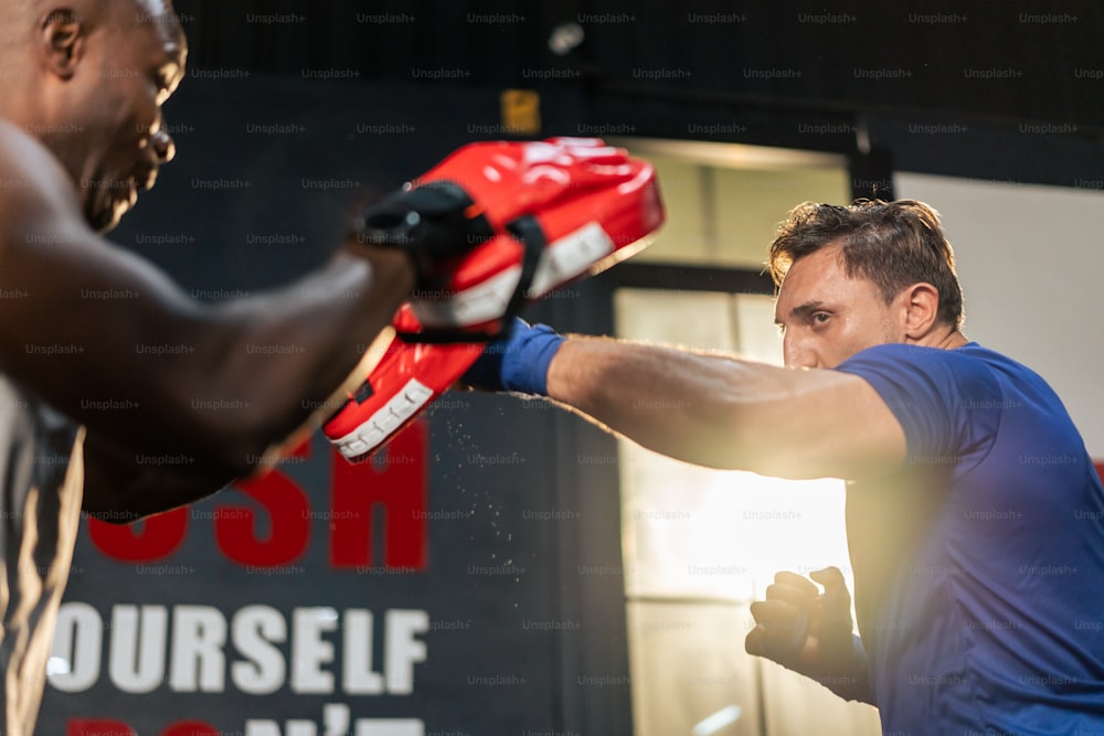 Kaukasier tragen Boxhandschuhe und schlagen mit afrikanischem Trainer voraus. Attraktiver junger Bodybuilder, der mit Hilfe eines afroamerikanischen Trainers für die Gesundheitsversorgung im Fitnessstudio oder Fitnessclub trainiert.