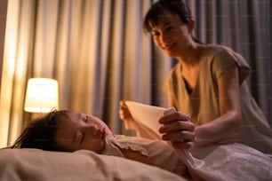 Asian loving beautiful mother take care asleep young small comfortable kid. Caring parent mom put blanket on sleeping little girl daughter on bed in dark bedroom at night. Parenting activity at home.