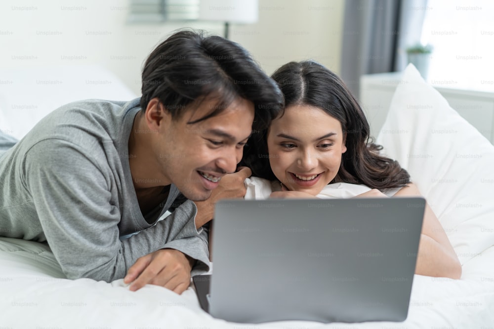 Asian attractive happy young couple watching movie on bed in bedroom. Beautiful new marriage man and woman in pajamas feel happy and relax in early morning after wake up use laptop computer at home.