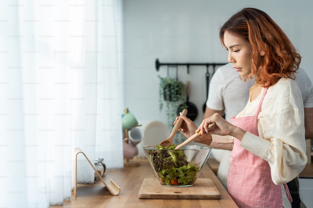 아시아의 아름다운 여자는 집에서 부엌에서 남편과 함께 그린 샐러드를 요리합니다. 젊은 매력적인 부부는 행복을 느끼고 야채를 먹는 것을 즐깁니다. 건강 식품을 다이어트하고 집에서 건강 관리를 위해 체중을 줄입니다.