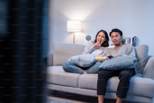 Asian loving young couple watch movie together in living room at home. Attractive new marriage man and woman sit on sofa, using remote control TV show and have fun laughing look to television in house