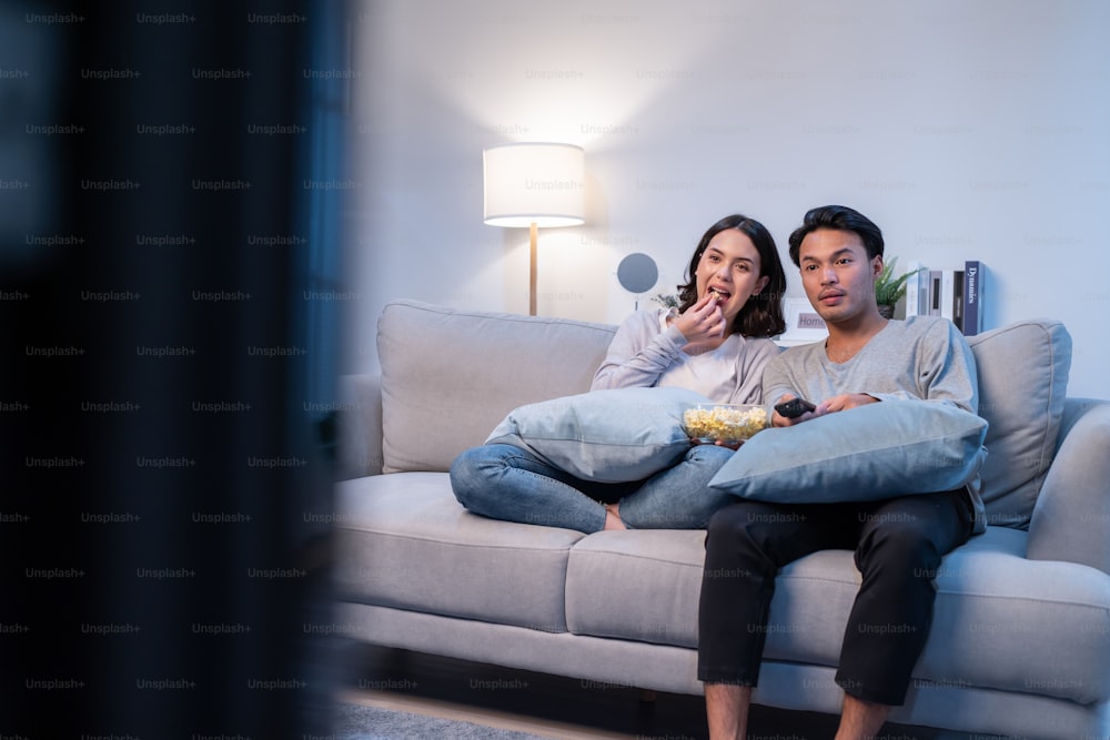 Asian loving young couple watch movie together in living room at home. Attractive new marriage man and woman sit on sofa, using remote control TV show and have fun laughing look to television in house