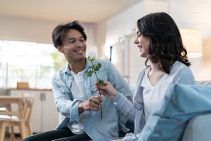 Asian young man surprise and give rose flower to beautiful girlfriend. Attractive romantic new marriage couple male and woman spend time together in living room at home, celebrate anniversary in house