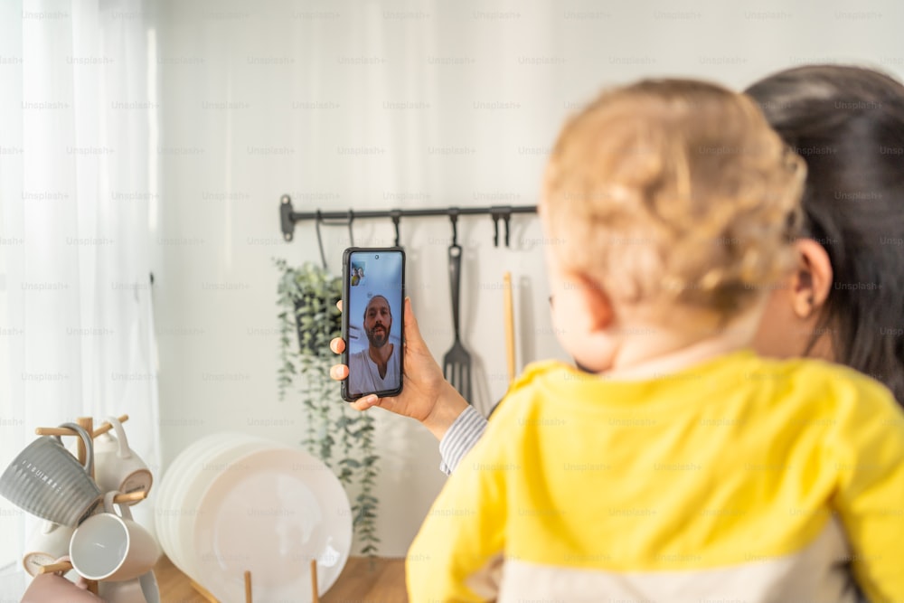 Caucasian mother holding little baby and video call online with family. Attractive beautiful mom using mobile cellphone and chat with husband while carrying young toddler kid in living room in house.