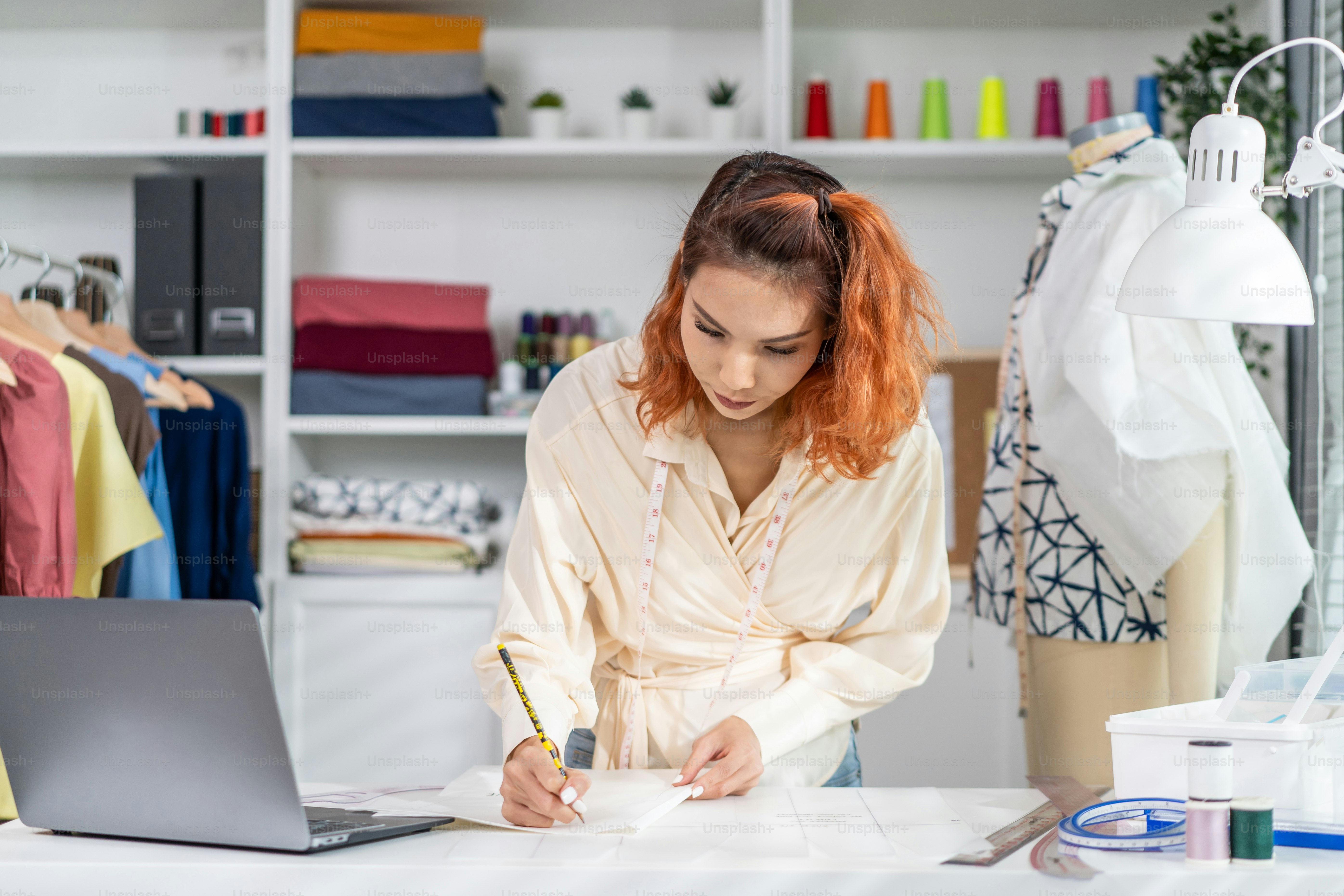 Asian beautiful tailor woman working on clothes in tailoring atelier. Attractive female fashion designer dressmaker design drawing and sketching out ideas for new fashion collection in workshop room.