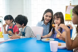 Asiatische schöne junge Lehrerin, die dem Kind im Schulzimmer einen Snack serviert. Attraktive Lehrermeisterin gibt dem Schüler eine Pause und serviert Kekse und Getränke an die Vorschule im Kindergarten im Klassenzimmer.