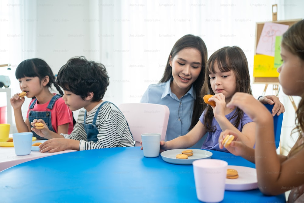 Asiatische schöne junge Lehrerin, die dem Kind im Schulzimmer einen Snack serviert. Attraktive Lehrermeisterin gibt dem Schüler eine Pause und serviert Kekse und Getränke an die Vorschule im Kindergarten im Klassenzimmer.