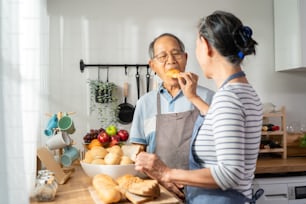 Asian Senior older couple spending time together in kitchen at home. Attractive man and woman enjoy cook foods for breakfast, grandmother feed bread to grandmother with happiness. Family relationship.