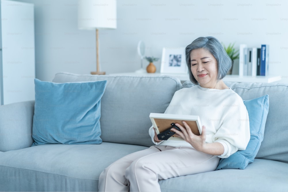 Asian Senior woman looks family photographs with gentle in living room. Loving beautiful elderly older female grandmother sitting on sofa enjoy retirement life, missing husband and relative in house.
