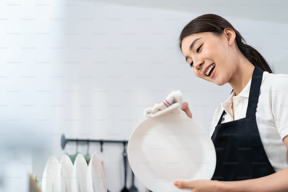 Asian active cleaning service woman worker cleaning in kitchen at home. Beautiful young girl housekeeper cleaner feel happy and wipes dishes plates after washing for housekeeping housework or chores.