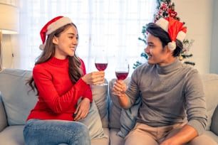 アジアの若い幸せなカップルは、家で一緒にクリスマスパーティーを祝います。魅力的な美しい男性と女性は、赤ワインのアルコールを飲み、ソファでグラスを乾杯し、自宅でクリスマスに幸せで興奮しています。