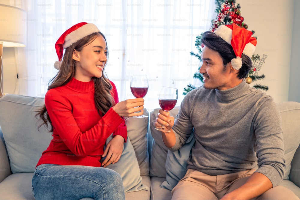 Asian young happy couple celebrate christmas party together in house. Attractive beautiful man and woman drinking red wine alcohol and toasting glass on sofa, feel happy and excited for Xmas at home.