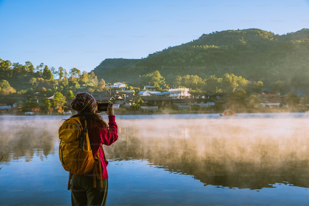 La donna in piedi vicino al lago, sorrideva, felice e si godeva la bellezza naturale della nebbia. Usa un telefono cellulare per scattare una foto della nebbia che galleggia sulla superficie del lago.