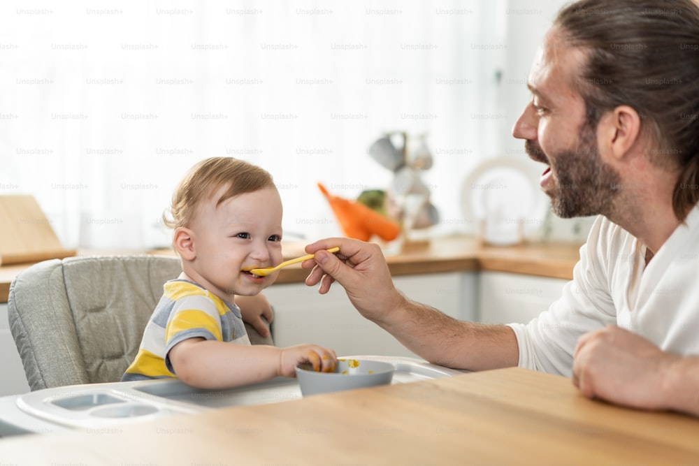 Caucasian beautiful father take care of baby boy toddler in kitchen. Happy family, Attractive loving dad cook and feeding healthy foods to little kid son infant while sit on chair for lunch in house.