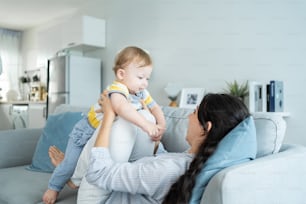 Caucasian loving mom play with baby boy child on sofa in living room. Happy family, attractive beautiful caring young mother lifting toddler son fly up enjoy doing airplane game in morning in house.