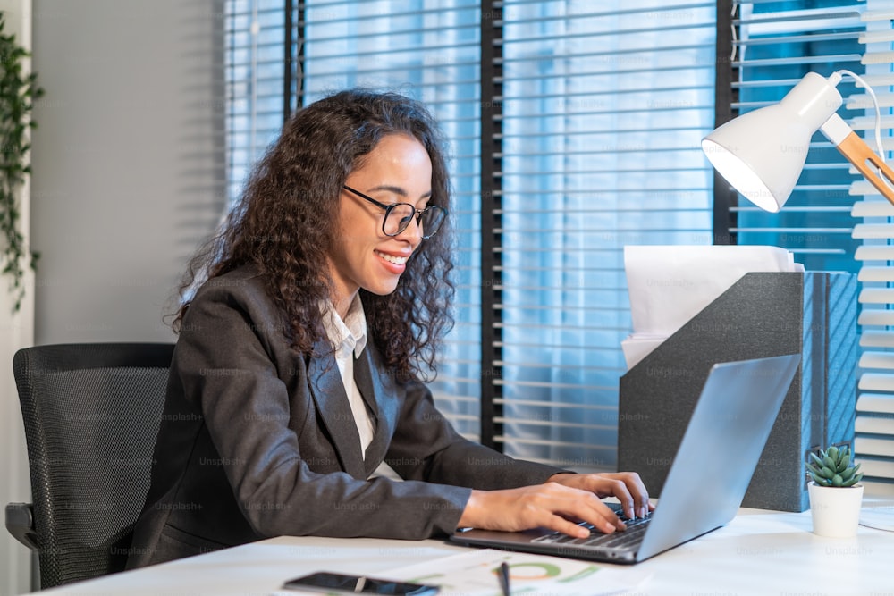 Happy beautiful Latino businesswoman in formal suit work in workplace. Attractive female employee office worker smile, use laptop computer for discuss with co worker and typing on keyboard in company