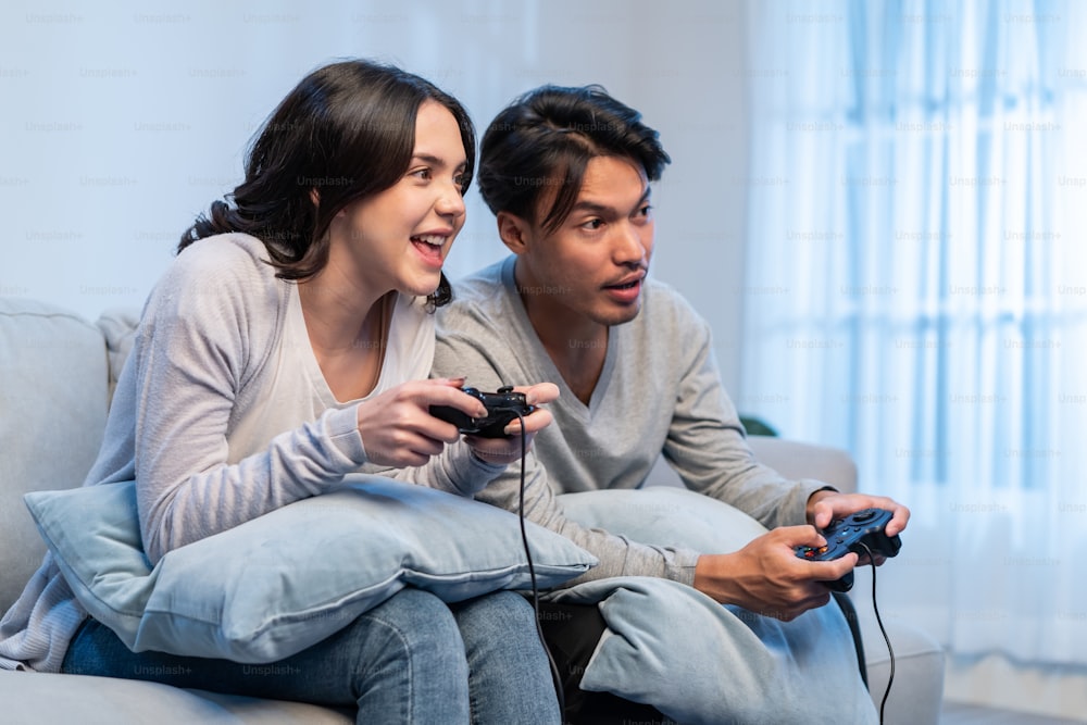 Asian young man and woman couple enjoy playing game together in house. Attractive funny male and female friend feeling happy sit on sofa in living room, spend time together play joystick game at home.