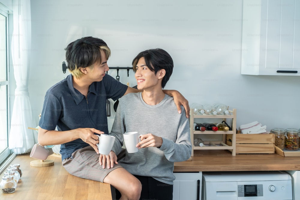 Asian man gay family sitting on counter, drink coffee for breakfast. Attractive romantic male lgbtq couple celebrate anniversary, spend time have dinner in kitchen in house. Homosexual-LGBTQ concept.