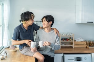 Asian man gay family sitting on counter, drink coffee for breakfast. Attractive romantic male lgbtq couple celebrate anniversary, spend time have dinner in kitchen in house. Homosexual-LGBTQ concept.