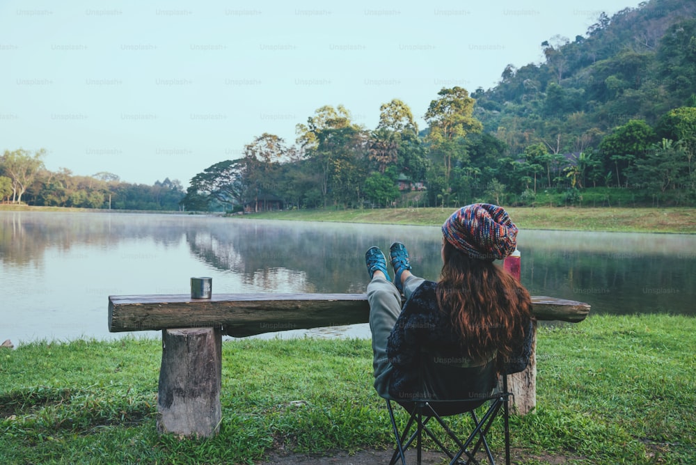 アジアの女性は、木製のテーブルに足を乗せて、湖畔でモーニングコーヒーを楽しむことでリラックスできます。キャンプ旅行