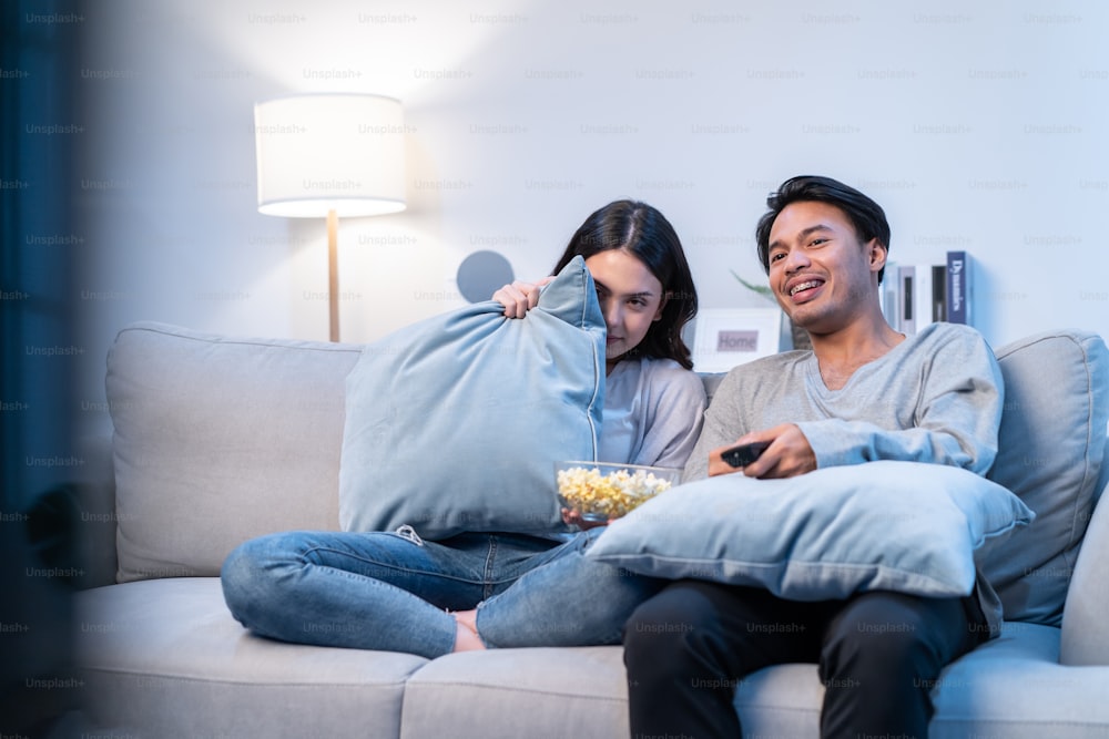 Asian young loving couple watch movie together in living room at home. Attractive man and woman sit on sofa, boyfriend with girlfriend spend time watch scary jumpscare on TV in living room in house.
