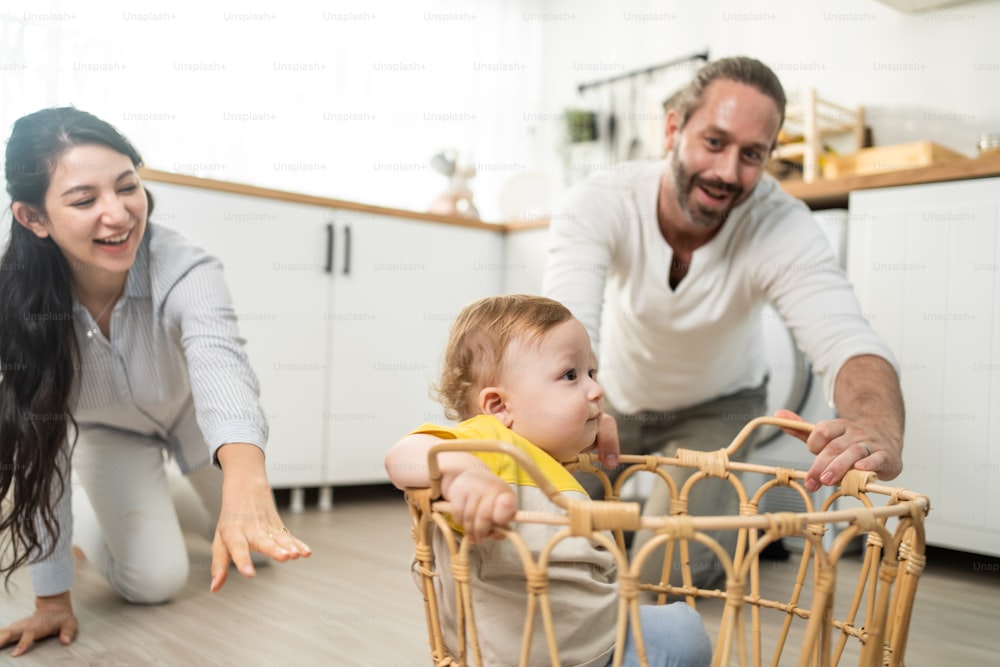 Caucasian loving parents play with baby toddler in kitchen on floor. Beautiful couple mother and father play with young little son child and put infant in basket. Activity relationship at home concept