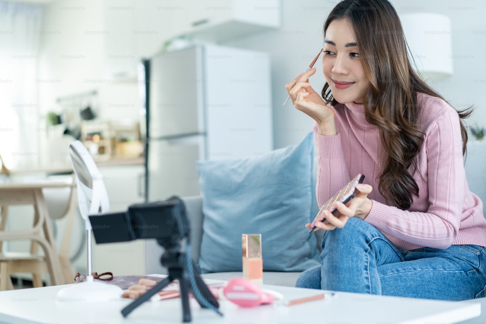 Vlogger fêmea asiática conversa com câmera para rever o cosmético em casa. Bela blogueira de beleza Influencer menina sentar-se no sofá e transmissão ao vivo, mostrando paletas de sombras maquiagem para os seguidores em casa.