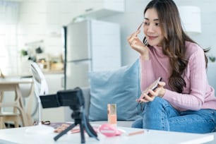 Las vloggas asiáticas hablan con la cámara para revisar el cosmético en casa. Hermosa bloguera de belleza Chica influencer sentada en el sofá y transmitiendo en vivo, mostrando paletas de sombras de ojos a sus seguidores en casa.