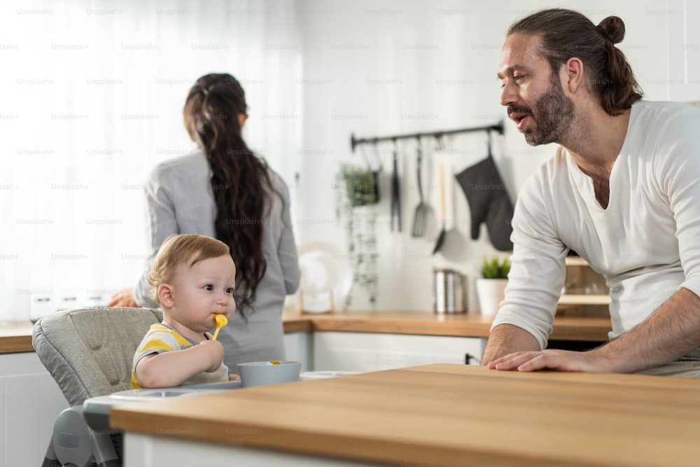 Caucasian beautiful parents take care of baby boy toddler in kitchen. Happy family, Attractive young couple cook and feed healthy foods to little kid son infant while sit on chair for lunch in house.