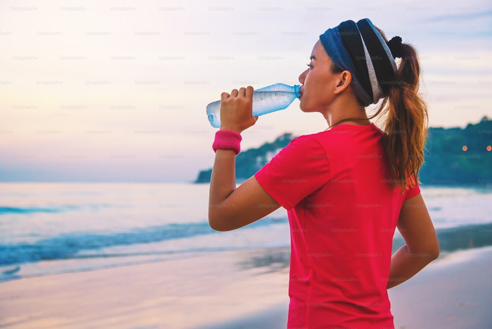 朝、ビーチでジョギングをするアジアの女性。海の散歩とペットボトルからの飲料水でリラックス