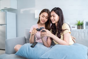 Dos amigas asiáticas disfrutan de las compras en línea en la sala de estar de casa. Hermosa pareja lesbiana se sienta en el sofá, sosteniendo la tarjeta de crédito para hacer el pago en línea del banco electrónico después de usar los productos de compra del teléfono en la tienda web