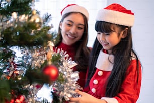 Asian lovely family, mother and kid daughter decorating Christmas tree.  Young little daughter feeling happy and excited to celebrate holiday Christmas Thanksgiving party together with parents in hous