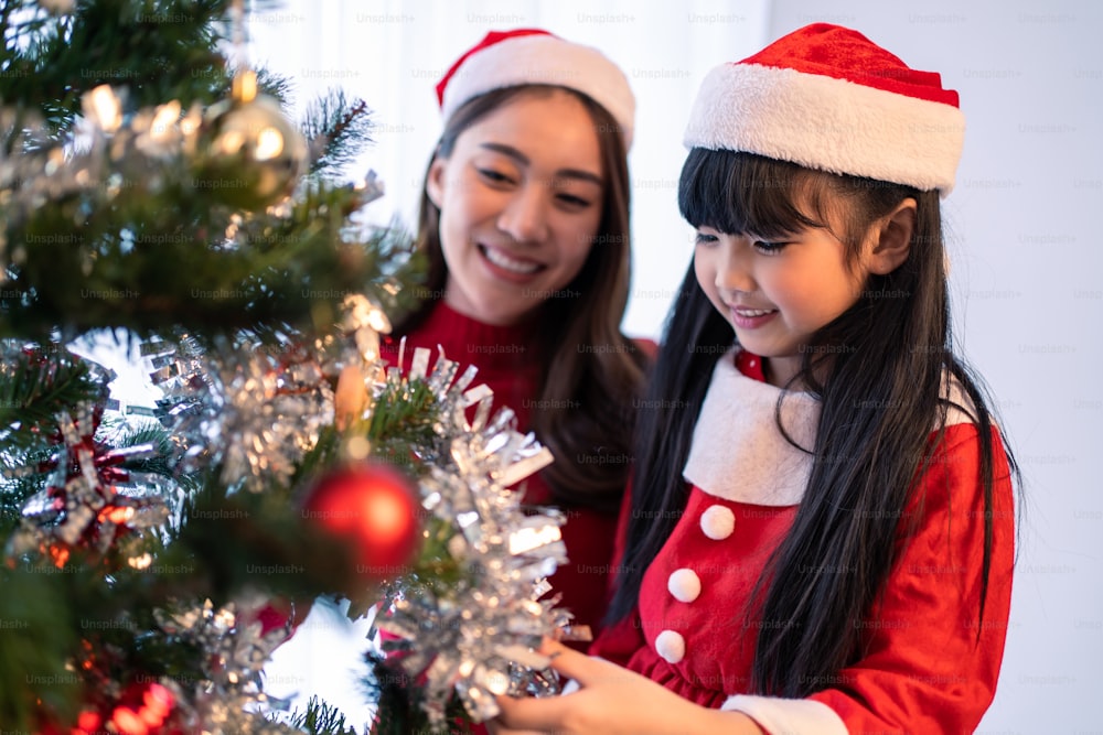 Bella famiglia asiatica, madre e figlia del bambino che decorano l'albero di Natale.  Giovane figlioletta che si sente felice ed entusiasta di celebrare la festa di ringraziamento di Natale di festa insieme ai genitori in casa
